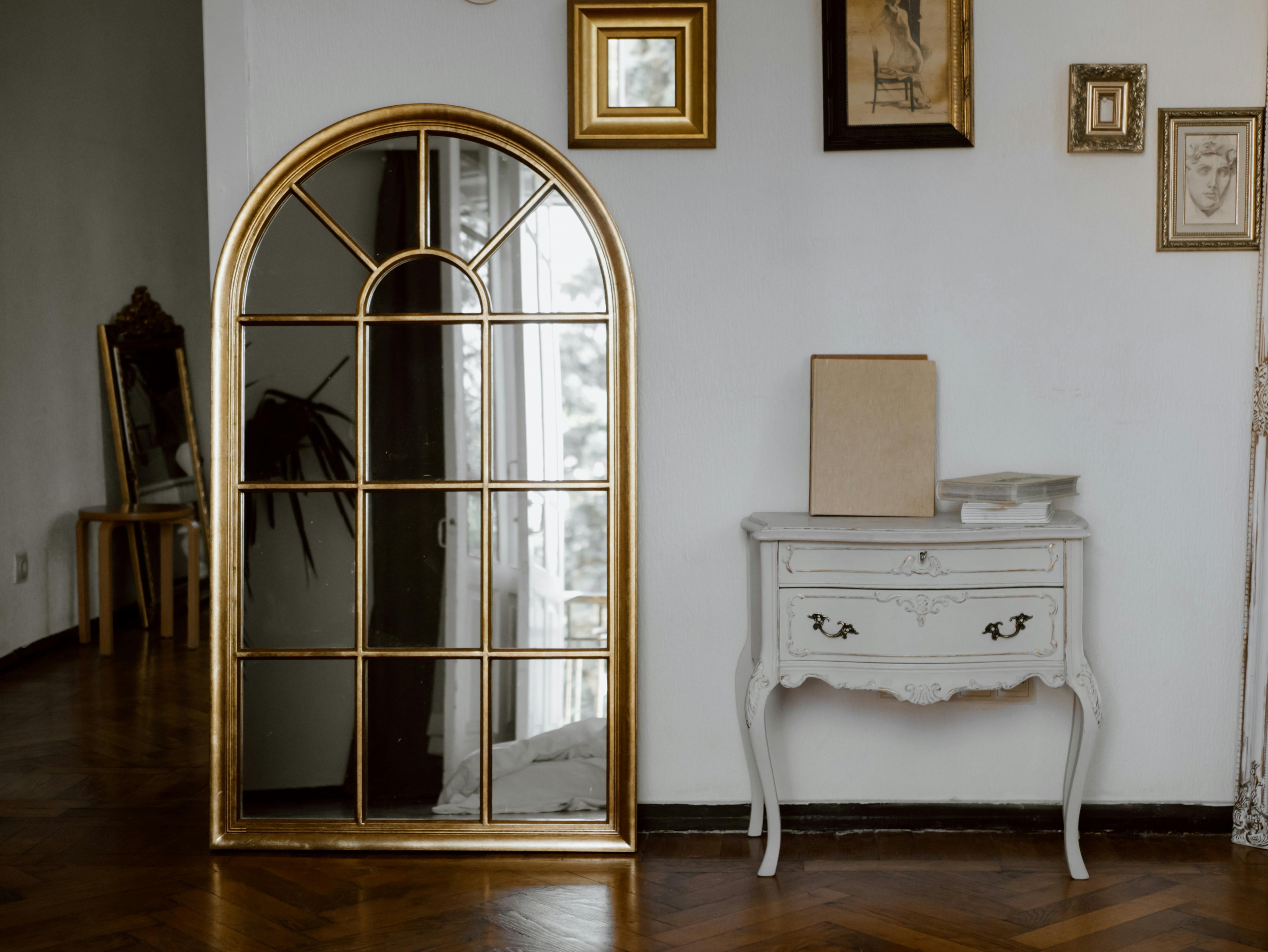 Vintage gold-framed arched mirror and distressed white dresser styled in a softly lit moody room.